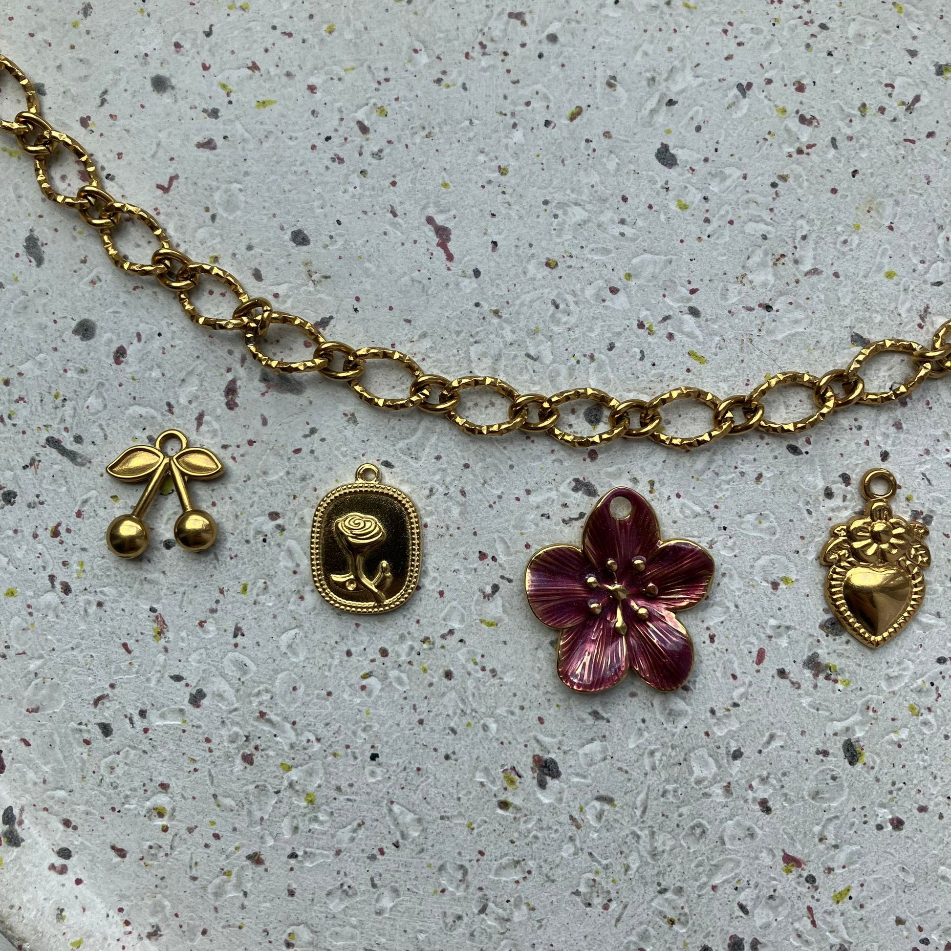 Close-up van een gouden ketting met bedels, van links naar rechts: een gouden kers, een rechthoekige bedel met een gedetailleerde roos, een grote bordeauxrode bloem, en een hartvormige bedel met een bloem erbovenop – allemaal uitgevoerd in goud | Zafia Jewellery
