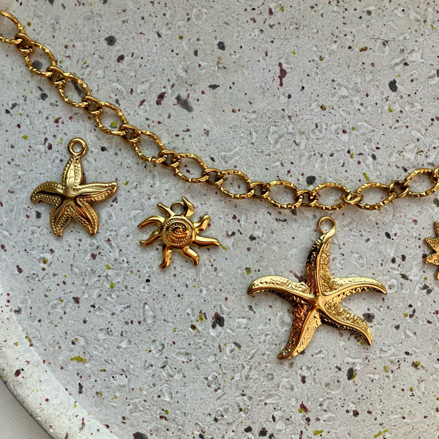 Close-up van drie gouden bedels: een gedetailleerde zeester, een zonnetje en een grote zeester.
