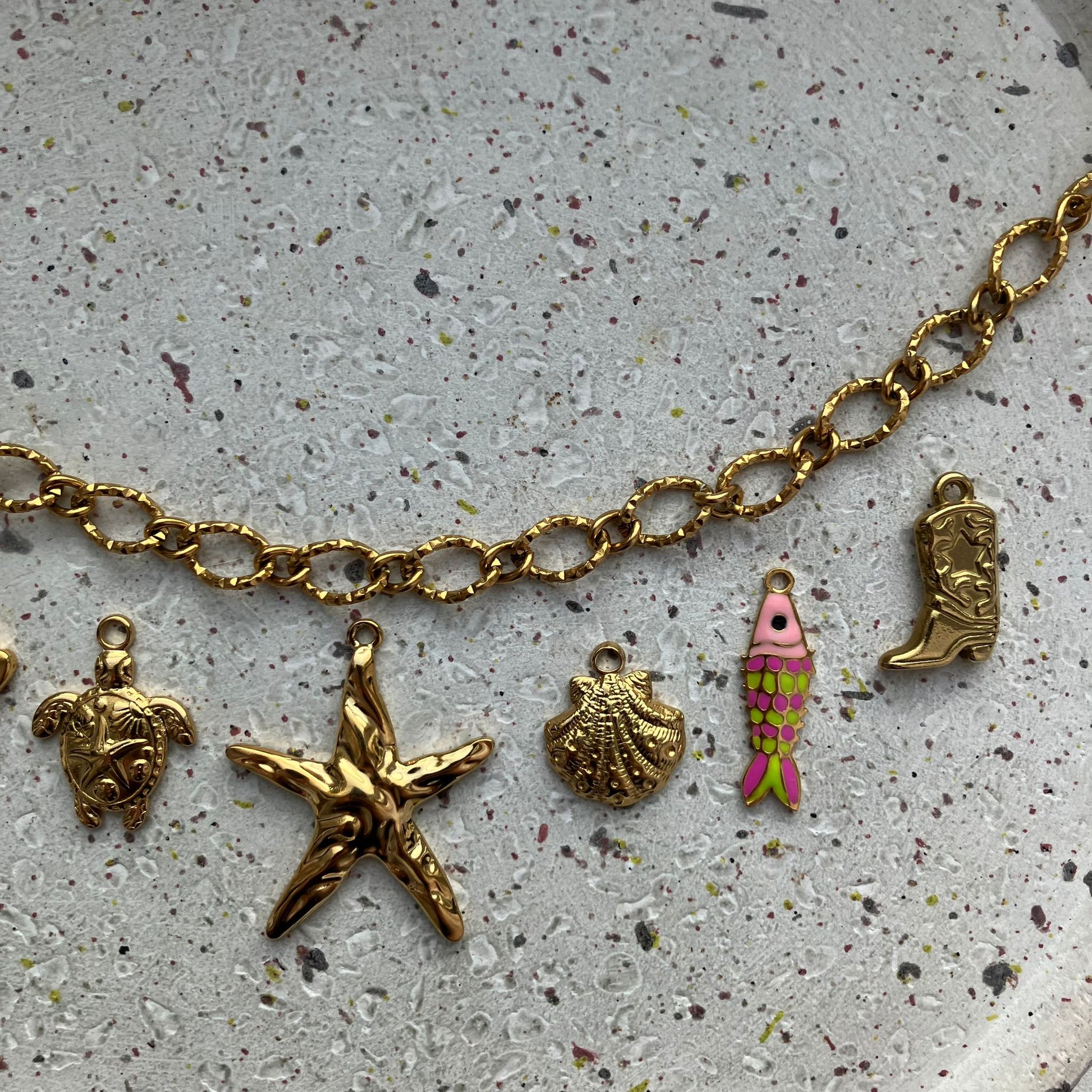 Close-up van gouden bedelketting met zomerse bedels: schildpad, zeester, schelp, visje met roze/groene schubben, cowboylaars | Zafia Jewellery