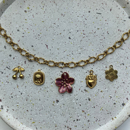 Een gouden, getextureerde ketting met vijf bedels van links naar rechts: een kersvormige bedel, een vierkant bedeltje met een roos erin, een grote bordeauxrode bloem met een gouden hart, een gedetailleerd hartvormig bedeltje met een bloemetje erbovenop, en een zonnebloem | Zafia Jewellery