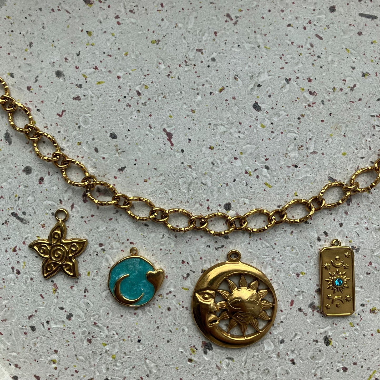 Gouden getextureerde ketting met vijf gouden bedels van links naar rechts: een gedetailleerde zon, een blauwe ronde bedel met een hartje als detail, een gecombineerde maan en zon, een rechthoekige bedel met een klein blauw steentje, en een ronde bedel met een zonmotief.