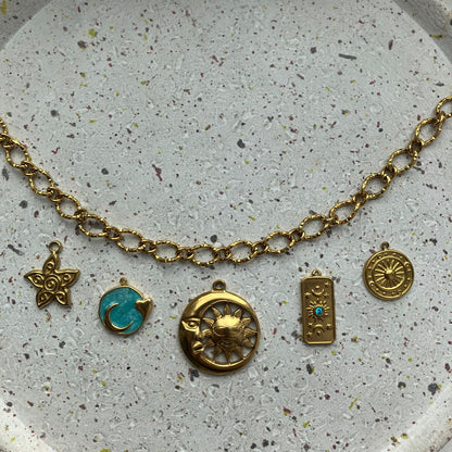 Gouden getextureerde ketting met vijf gouden bedels van links naar rechts: een gedetailleerde zon, een blauwe ronde bedel met een hartje als detail, een gecombineerde maan en zon, een rechthoekige bedel met een klein blauw steentje, en een ronde bedel met een zonmotief.