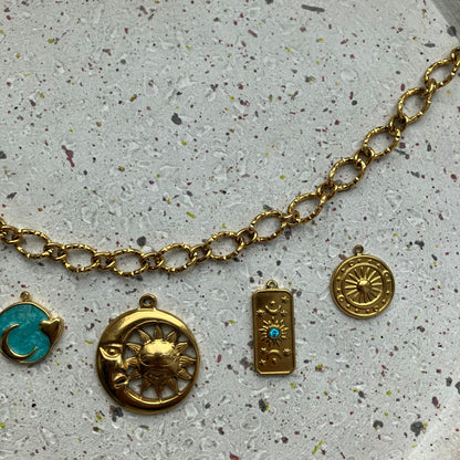 Close-up van een gouden getextureerde ketting van Zafia Jewellery met vijf bedels: zon, blauwe ronde met hartje, maan en zon, rechthoek met blauw steentje, en ronde zonbedel.